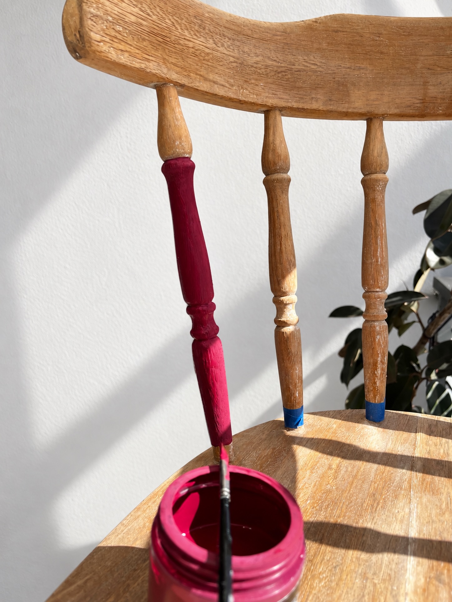 Pincel y frasco de pintura junto al respaldo de una silla de madera, con uno de los barrotes ya pintado en borravino y el resto sin pintar, planta de fondo